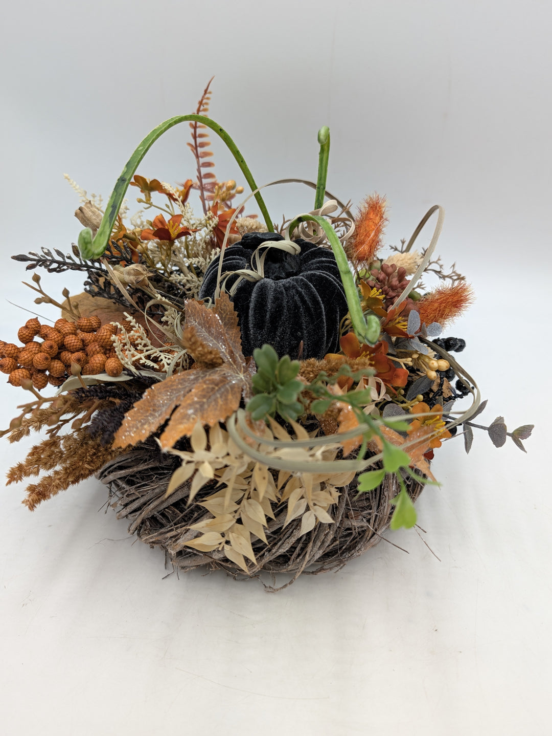 Herbstgesteck mit schwarzem Samtkürbis, orangefarbenen Blüten, Farn und Gräsern in rundem Reisignest