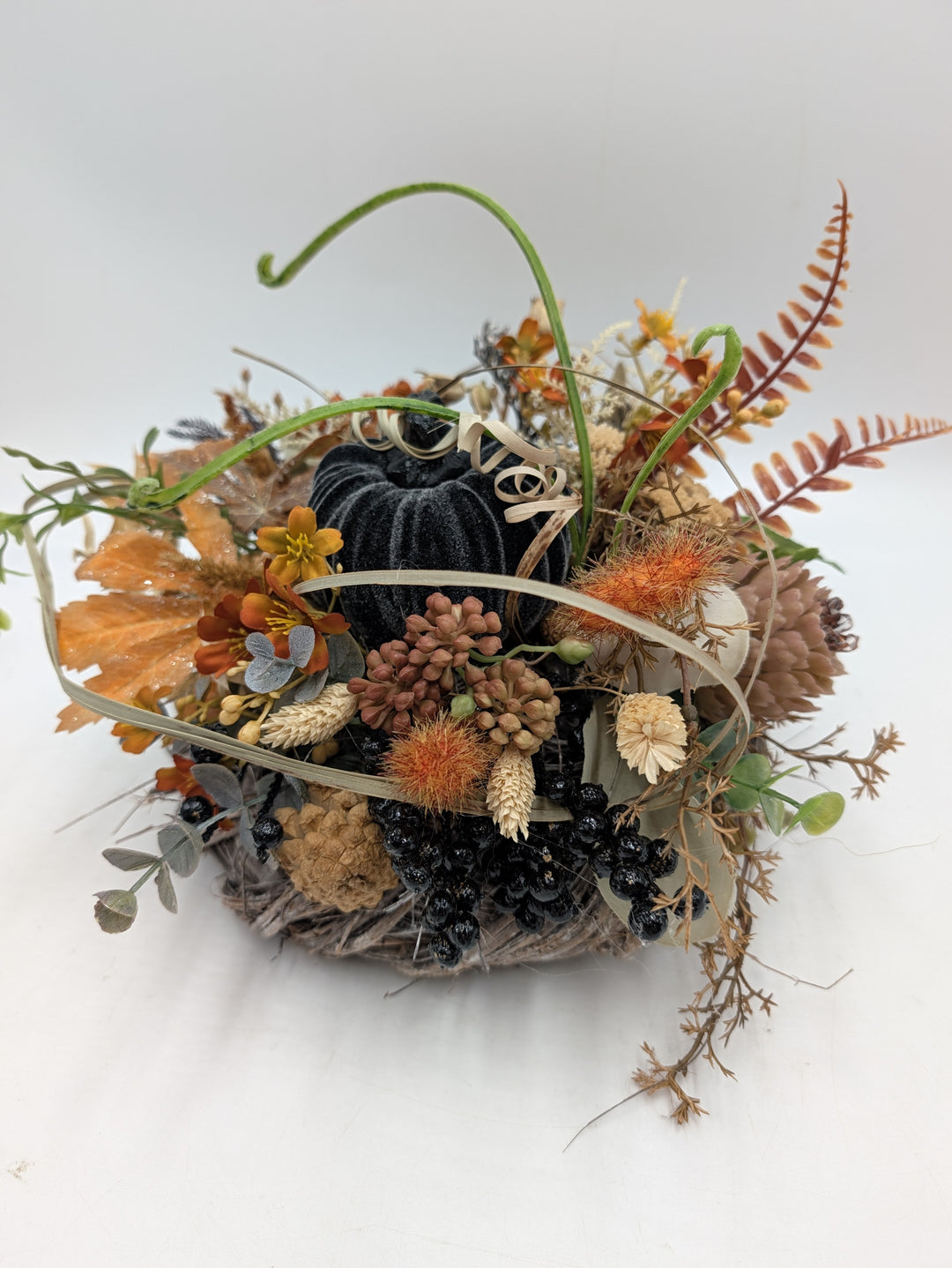 Aufsicht auf das Herbstgesteck mit schwarzen Beeren, Zapfen, Ähren und hellen Ranken um den Samtkürbis