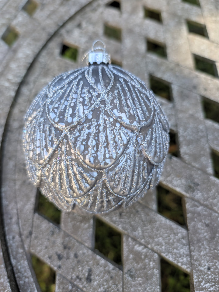 Silberne Christbaumkugel mit weißem Bogenmuster und Glitzer, hängend vor einem hellen Metallgeflecht