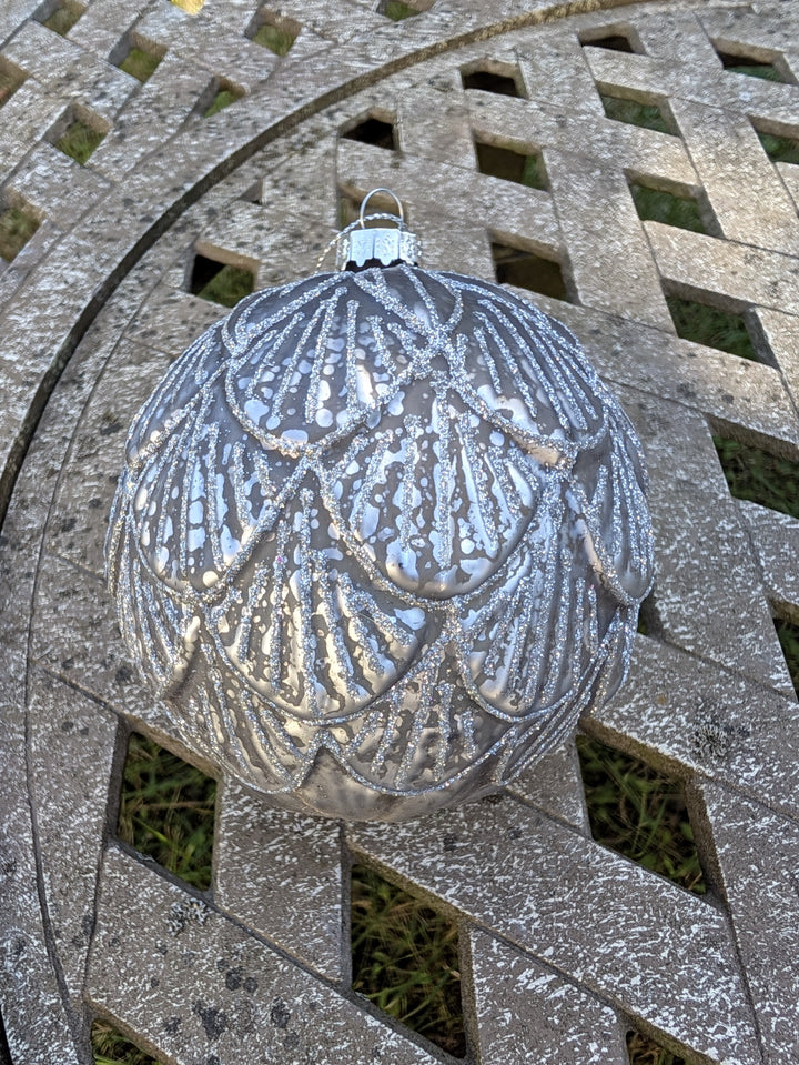 Weihnachtskugel in Silbergrau mit Glitzerbögen und heller Aufhängekappe, liegend auf einem Gartentisch