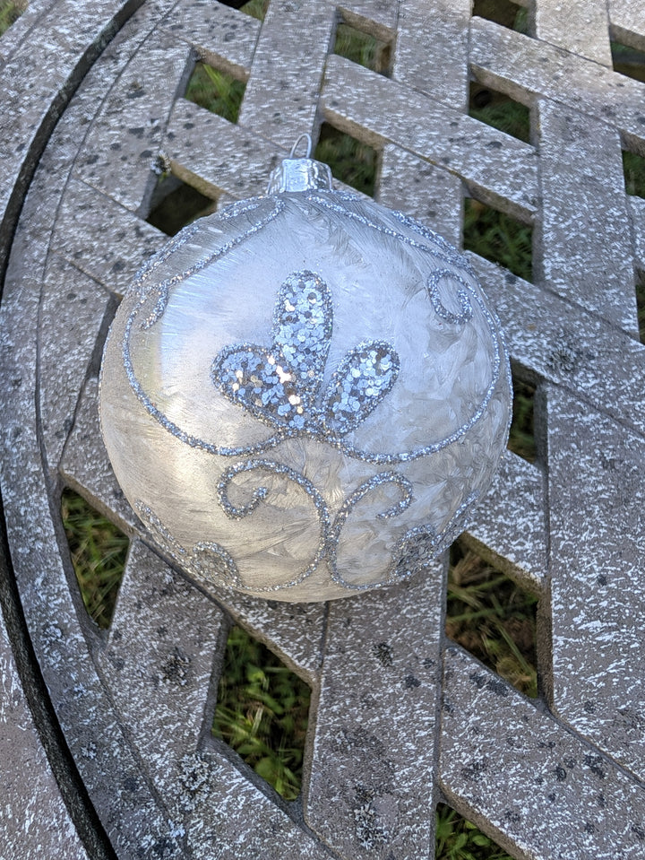 Silberweiße Christbaumkugel mit Glitzerranken und drei Paillettenblättern, liegend auf grauem Holzgitter