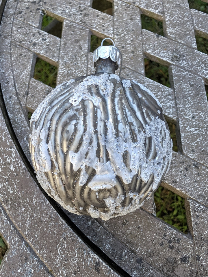 Christbaumkugel in Silber und Grau mit Rillenrelief und weißem Glitzer, liegend auf einem Gartentisch