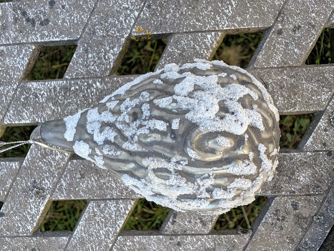 Tropfenförmiger Baumschmuck in Grau mit silbrigem Glitzerrelief und Aufhängeschlaufe auf Holzbank
