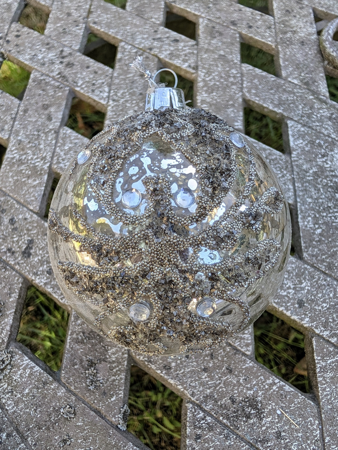 Klare Christbaumkugel mit silbernem Perlenmuster, Strasssteinen und Glitzerbesatz, hängend vor Holzlattenwand