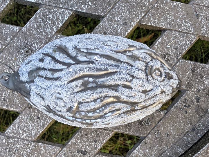 Tropfenförmiger Christbaumschmuck in Grau mit weißem Glitzer und Wellenrelief, liegend auf grauem Holzgitter
