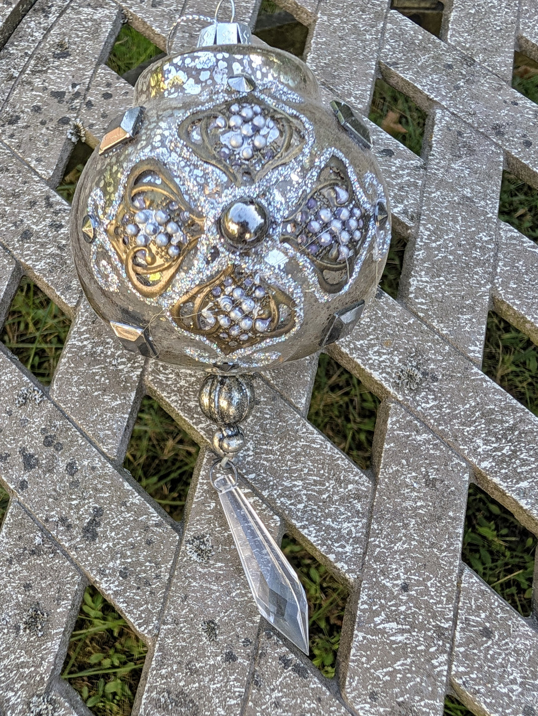 Silberne Christbaumkugel aus Glas mit goldenen Ornamenten, Perlen und klarem Anhänger, auf einer Holzbank