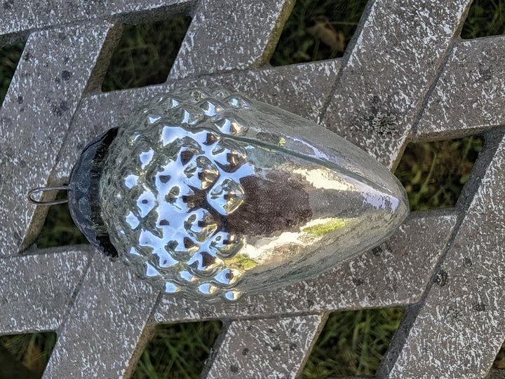 Silberner Zapfen aus Bauernglas mit Schuppenrelief, liegend auf einer hellen Holzbank