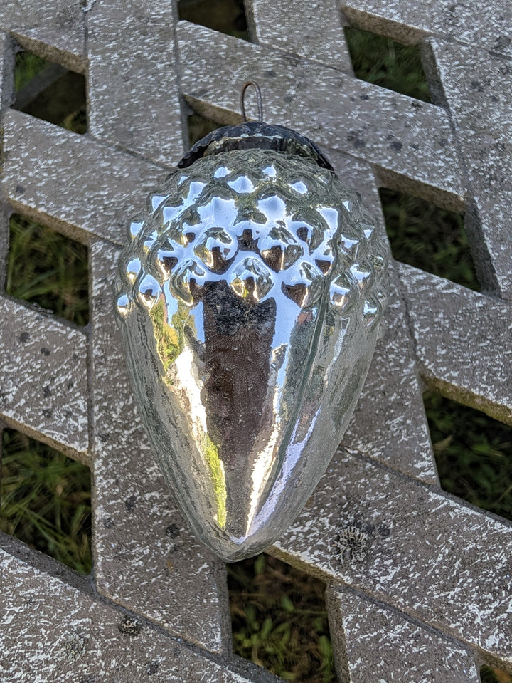 Glaszapfen in Silber mit Metallöse, hängend vor grauem Holzgitter, unten glatt und glänzend