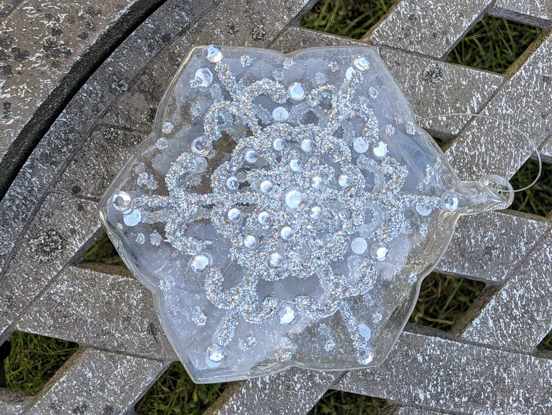 Klarer Glasstern in Schneeflockenform mit silbernem Glitzermuster und Strasssteinen, liegend auf grauer Holzbank