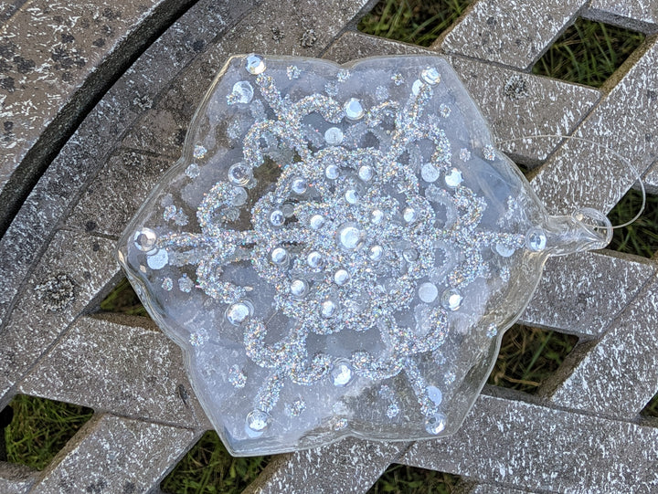Klarer Glasstern in Schneeflockenform mit silbernem Glitzermuster und Strasssteinen, liegend auf grauer Holzbank
