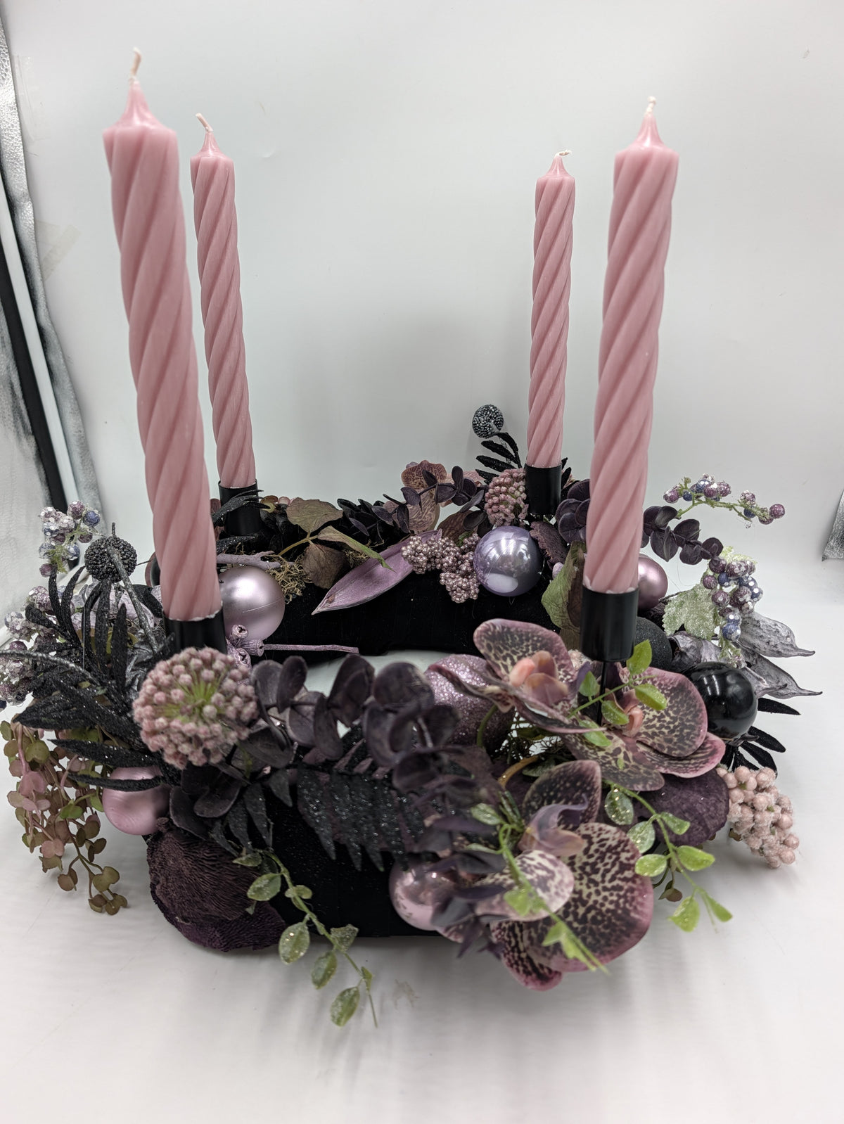 Adventskranz von der Seite: dunkle Eukalyptuszweige, schwarze Farnblätter, rosa Kugeln und vier Stabkerzen