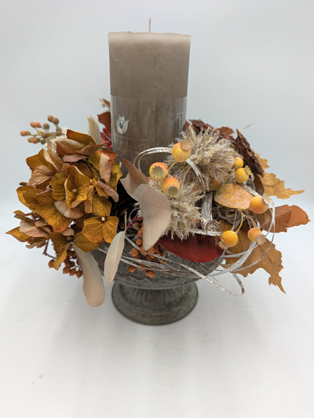 Herbstgesteck mit taupefarbener Stumpenkerze, orangebraunen Hortensien, Beeren und Herbstblättern in grauer Fußschale