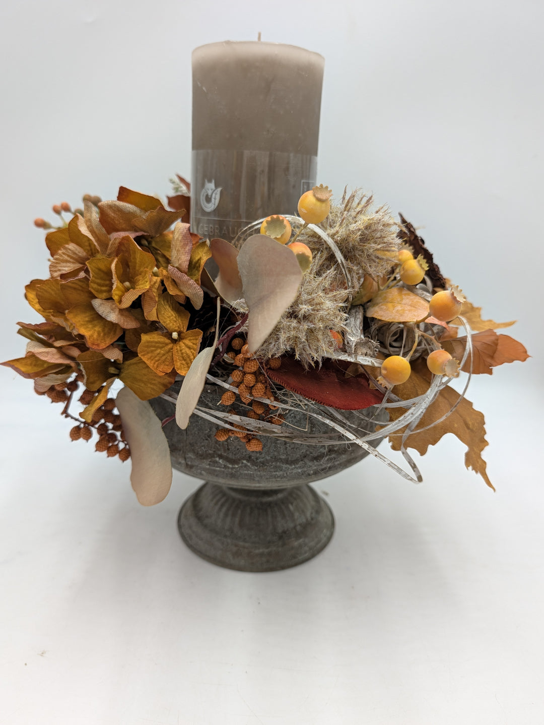 Herbstgesteck von der Seite: graue Amphorenschale mit Kerze, Hortensien, gelben Fruchtkugeln und hellen Gräsern