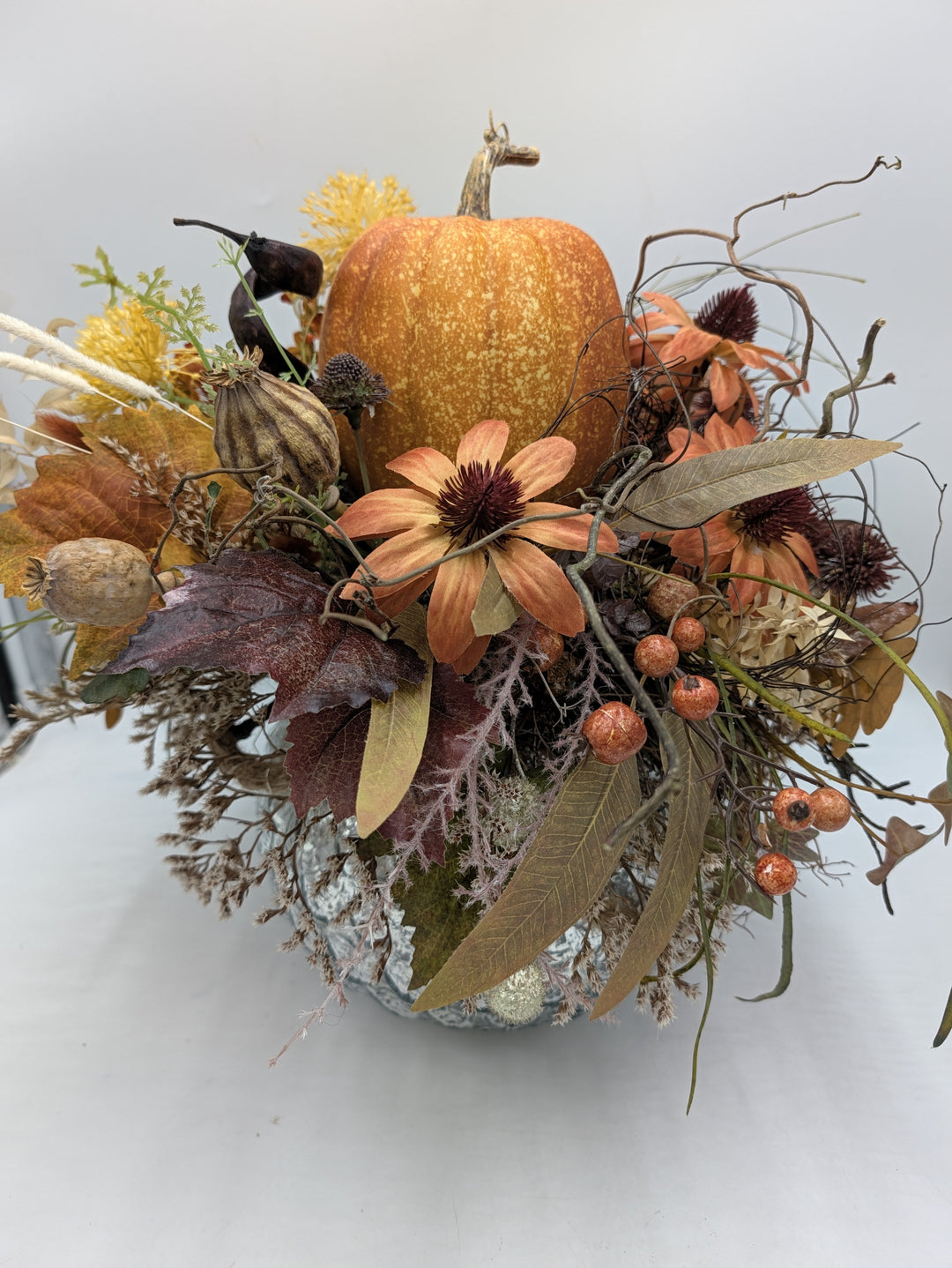 Herbstgesteck von vorn: Kürbis mit Stiel, orange Blüten, Mohnkapseln und Gräser in silbrig schimmernder Schale