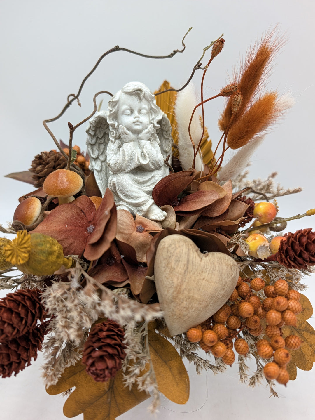 Nahaufnahme des Gestecks: Engelfigur, Holzherz, orangefarbene Beeren, Pilze und rostfarbene Gräser