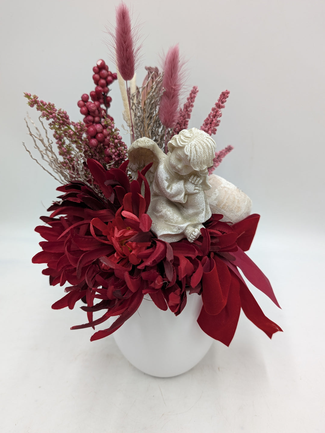 Grabgesteck von vorn: rote Chrysanthemen, rosa Beeren und Gr\u00e4ser, betender Engel auf hellem Stein