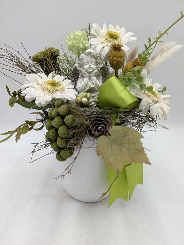 Trauergesteck mit grauer Engelfigur, weißen Gerbera, grünen Beeren und hellgrüner Schleife im weißen Topf