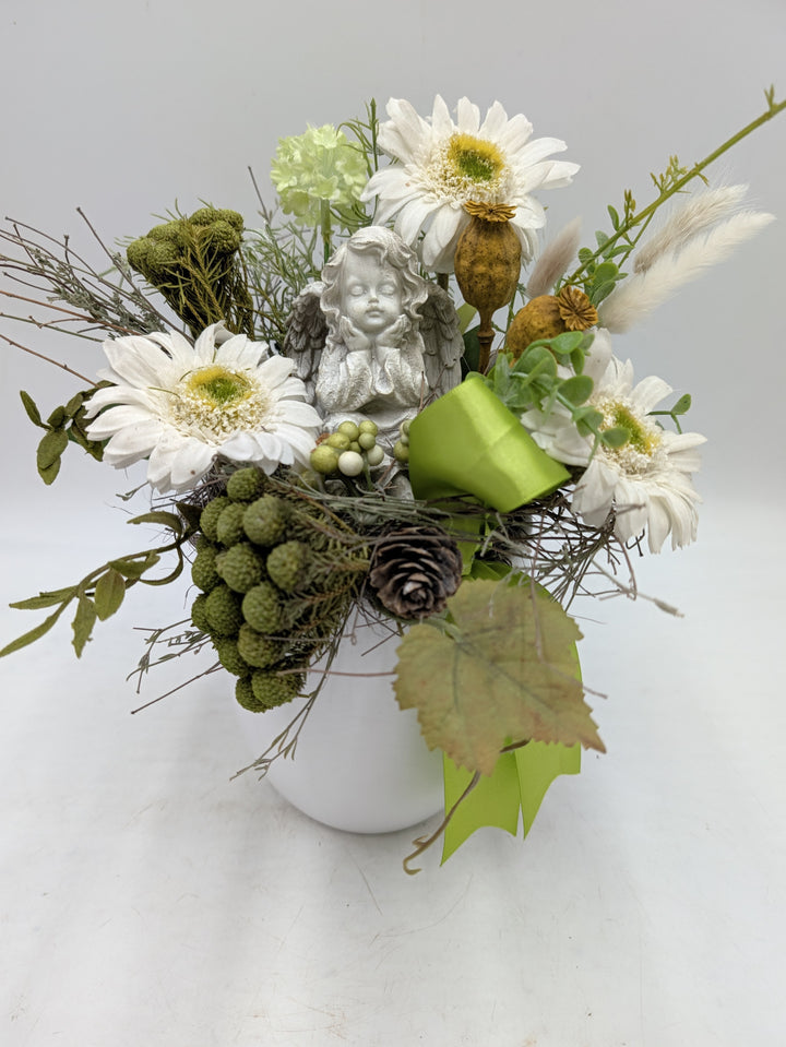 Trauergesteck von vorn: Engel zwischen weißen Gerbera, Mohnkapseln, Moos, Zapfen und grüner Satinschleife