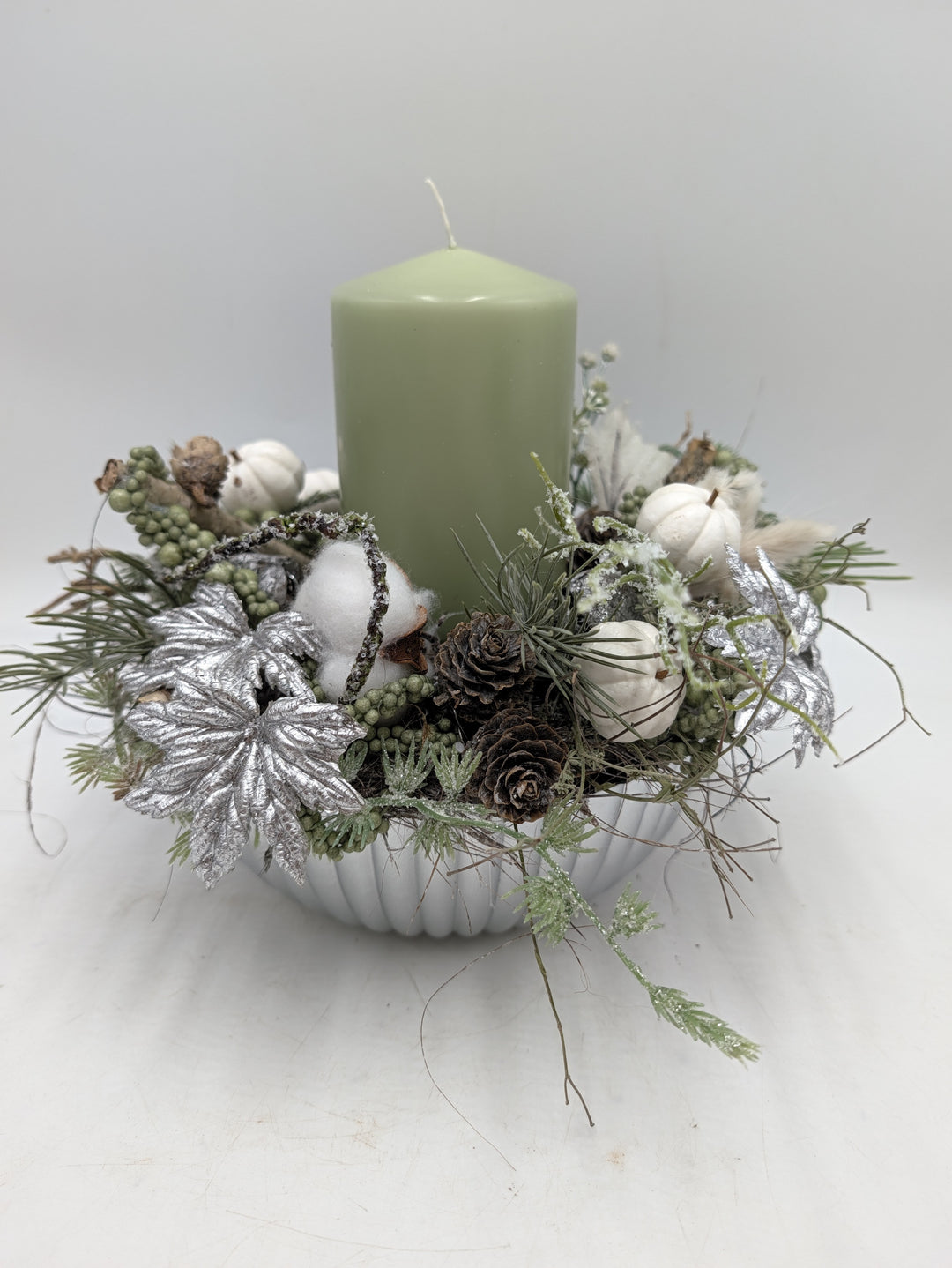 Wintergesteck mit mintgrüner Stumpenkerze, silbernen Blättern, Zapfen und Baumwolle in weißer geriffelter Schale