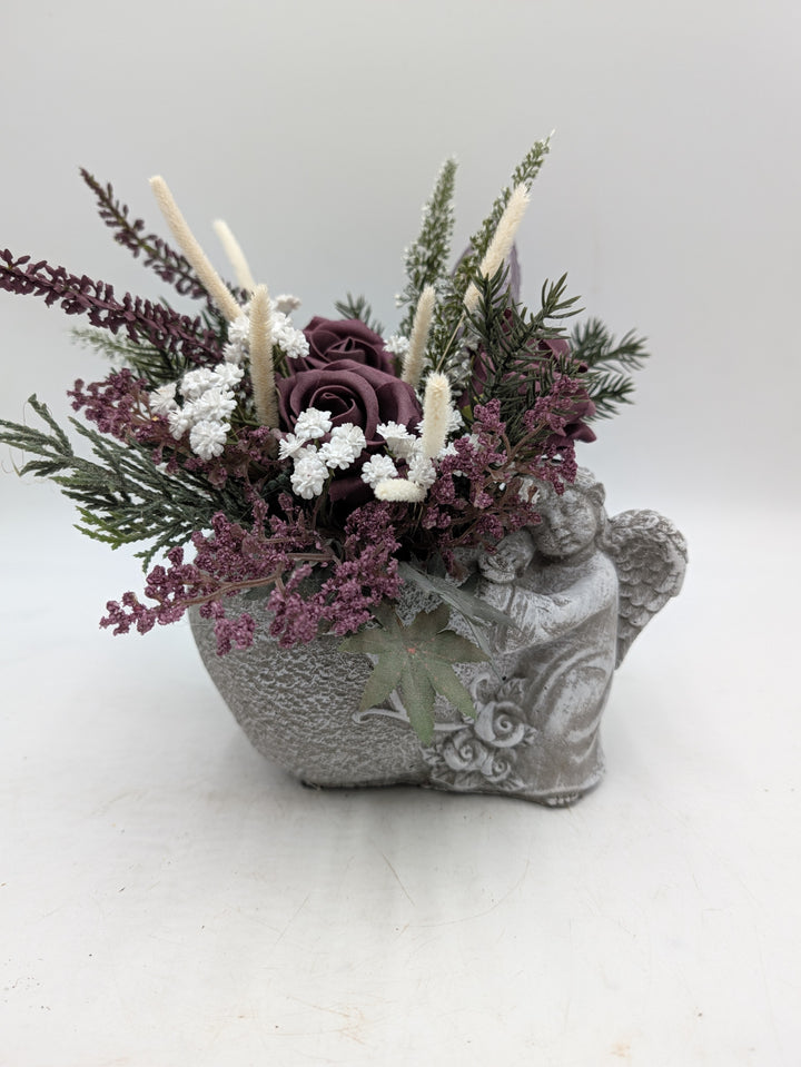 Trauergesteck mit lilafarbenen Rosen, weißen Blüten und Gräsern in grauem Gefäß mit Engelfigur, seitlich