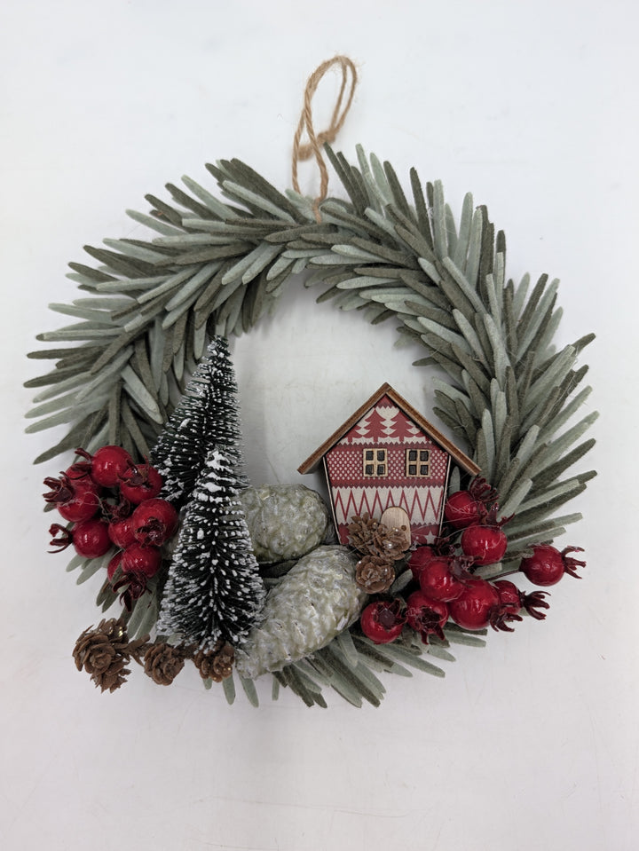 Weihnachtskranz mit graugrünen Nadeln, rotem Holzhaus, beschneiten Tannenbäumchen, roten Beeren und Zapfen
