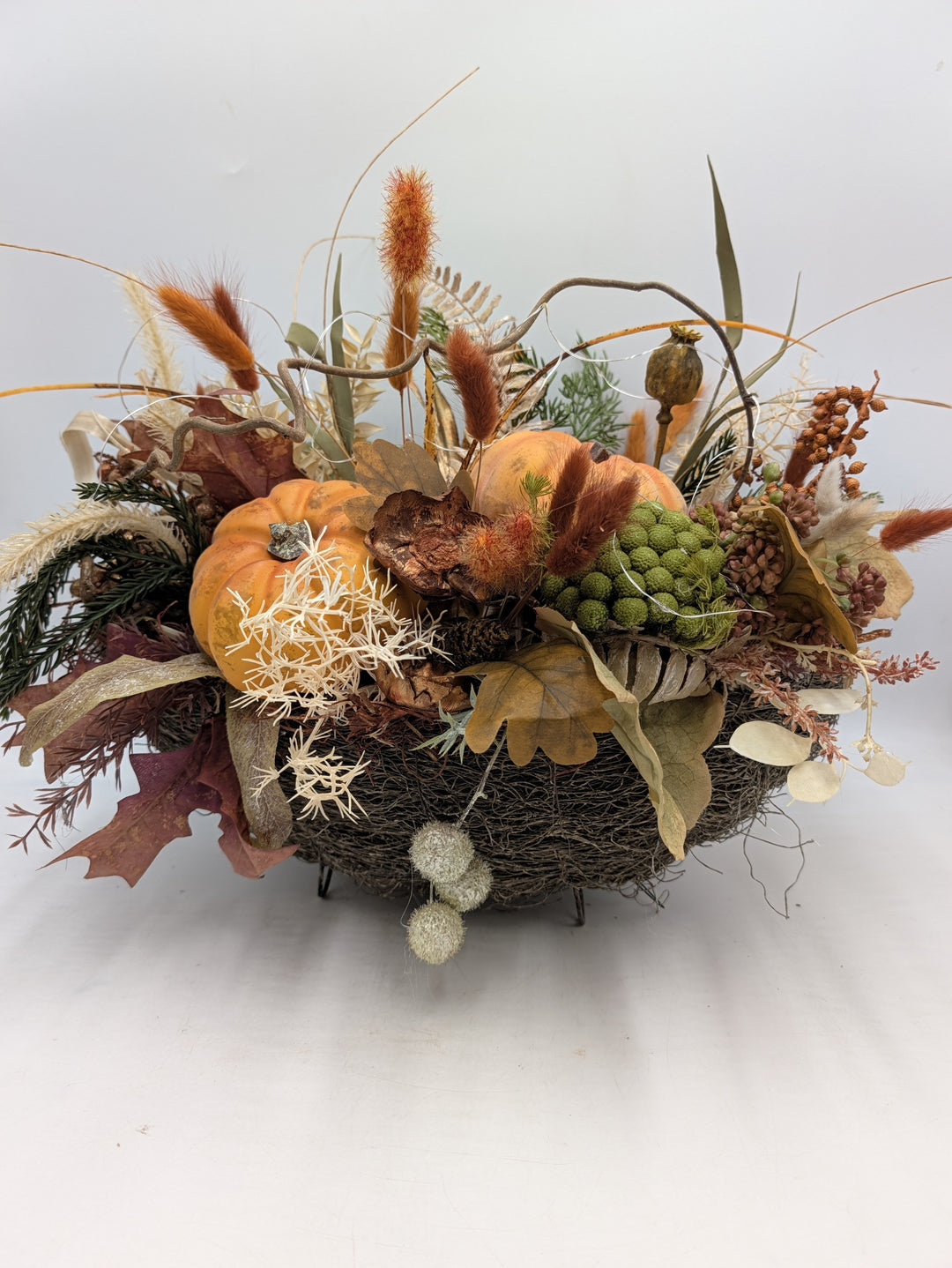 Herbstgesteck mit zwei orangefarbenen Kürbissen, Gräsern und Eichenblättern in flacher Reisigschale