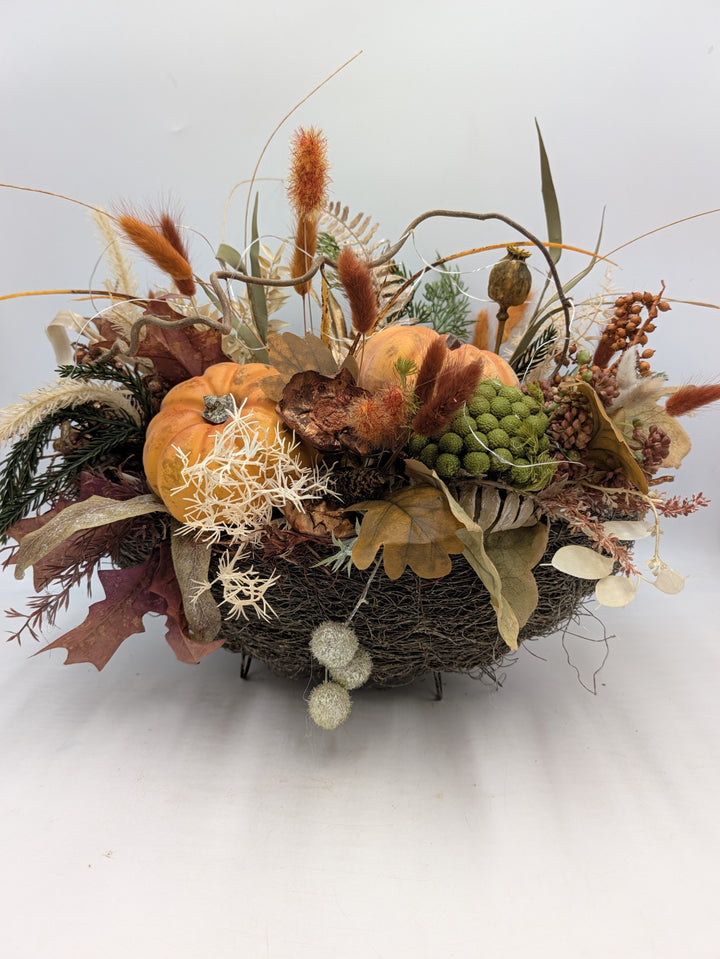Herbstgesteck mit zwei orangefarbenen Kürbissen, Gräsern und Eichenblättern in flacher Reisigschale