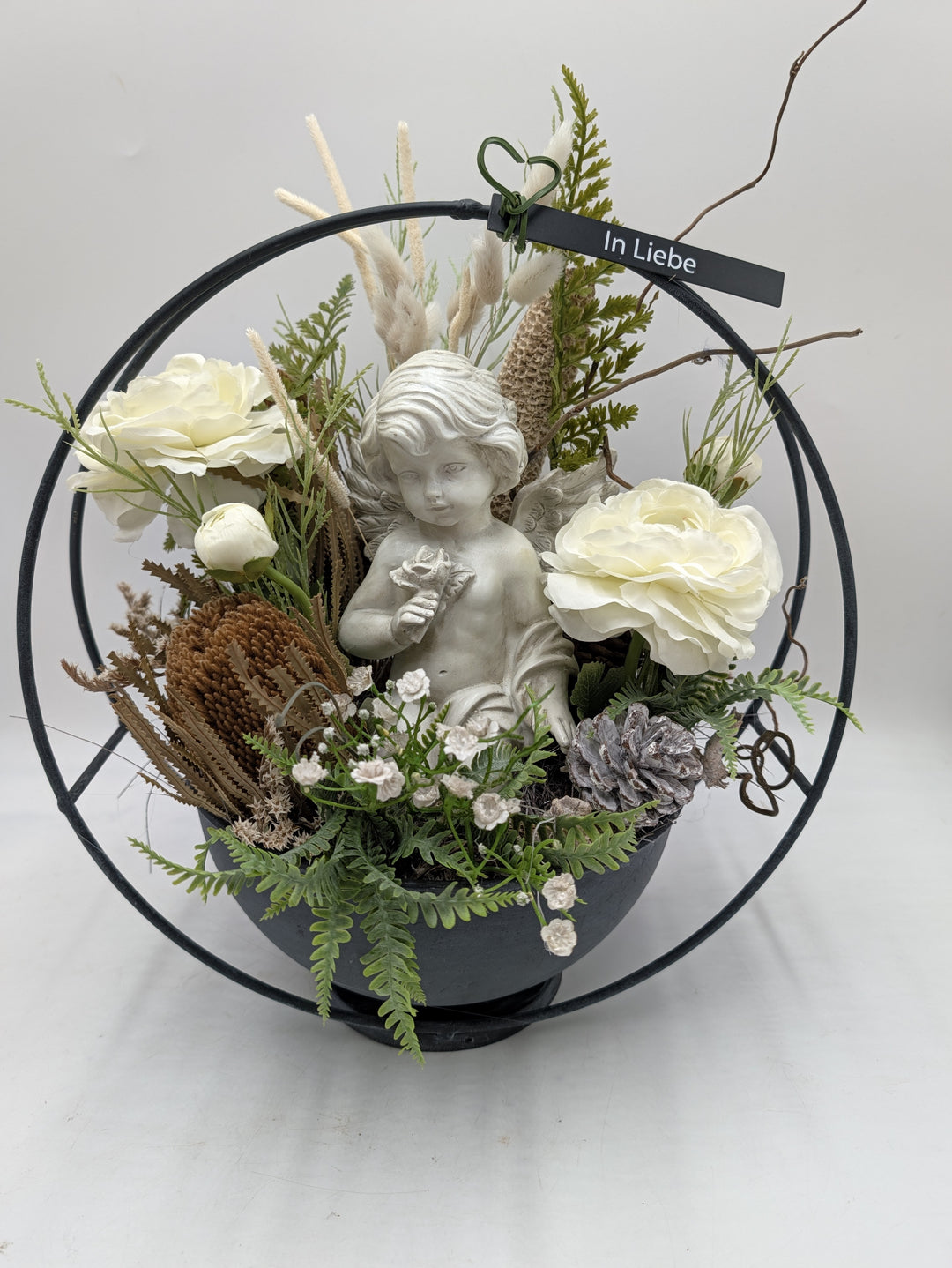 Trauergesteck mit Engelsfigur, weißen Rosen und Farn in grauer Schale im schwarzen Metallring, Schild In Liebe