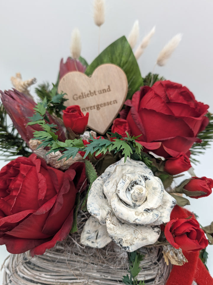Nahaufnahme: Holzherz mit Gravur „Geliebt und unvergessen“ zwischen roten Rosen, Protea und hellen Gräsern
