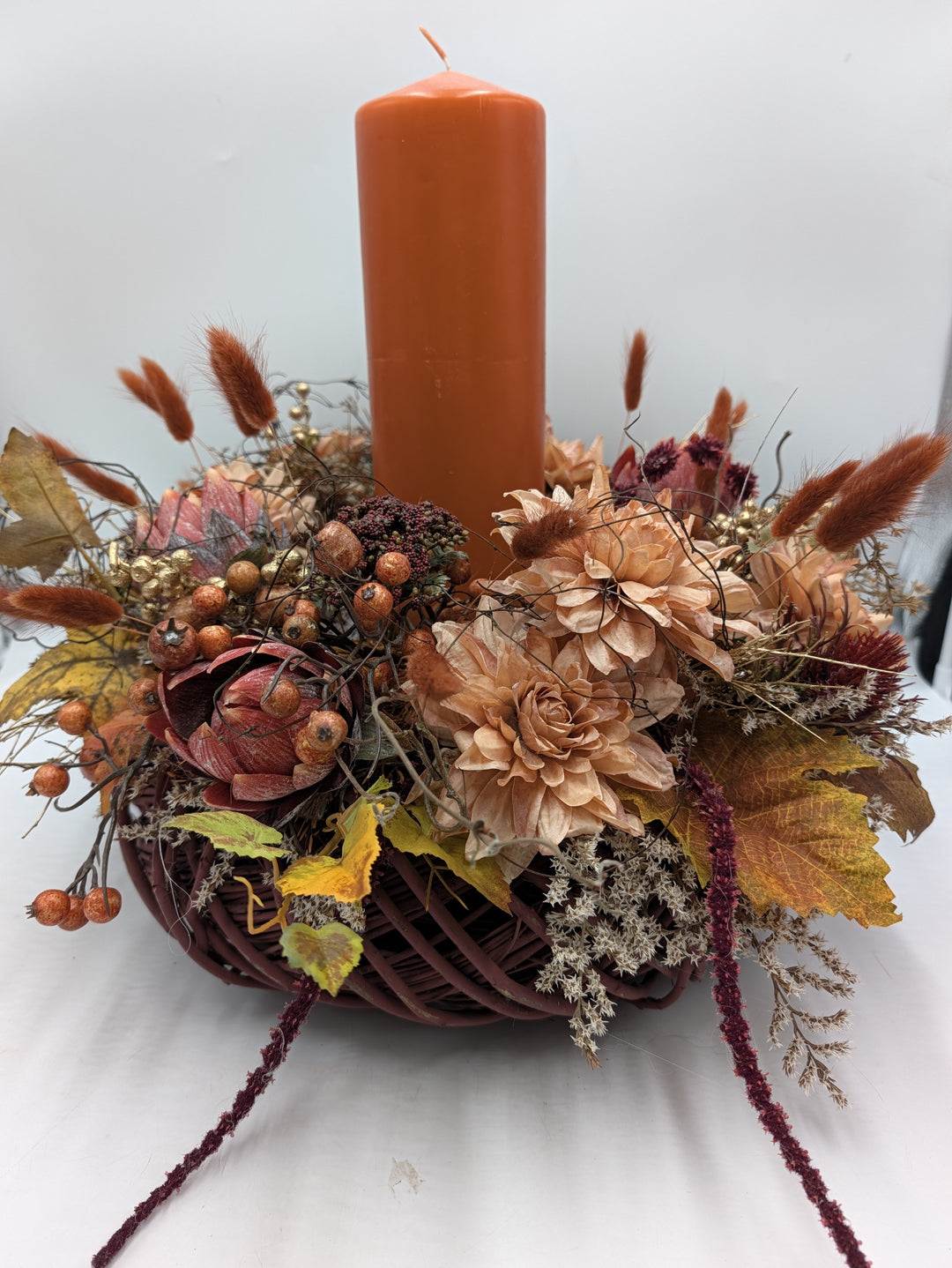 Herbstgesteck mit hoher orangefarbener Kerze, Dahlien in Apricot, Hagebutten und Gräsern in rundem dunkelrotem Korb