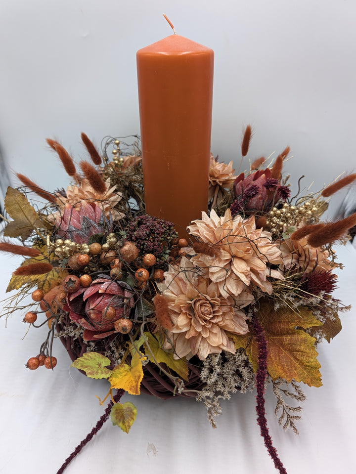 Herbstgesteck von vorn: orange Stumpenkerze, dunkelrote Proteen, goldene Beeren, Weinlaub und hängende Amarant-Rispen