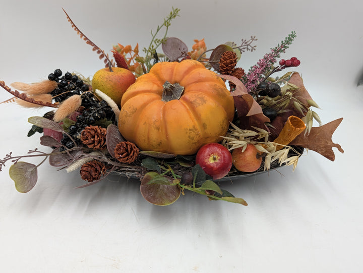 Flaches Herbstgesteck mit orangefarbenem Kürbis, Äpfeln, schwarzen Beeren, Zapfen und Heide in länglicher Schale