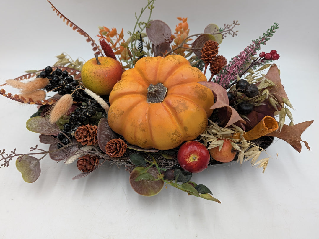 Herbstgesteck von vorn: Kürbis in der Mitte, umgeben von roten Äpfeln, Hafergräsern, Zapfen und rosa Heide
