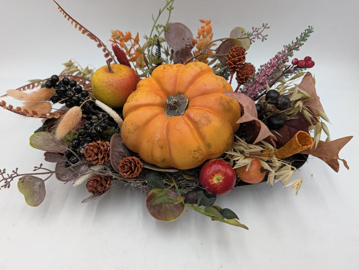 Herbstgesteck von vorn: Kürbis in der Mitte, umgeben von roten Äpfeln, Hafergräsern, Zapfen und rosa Heide