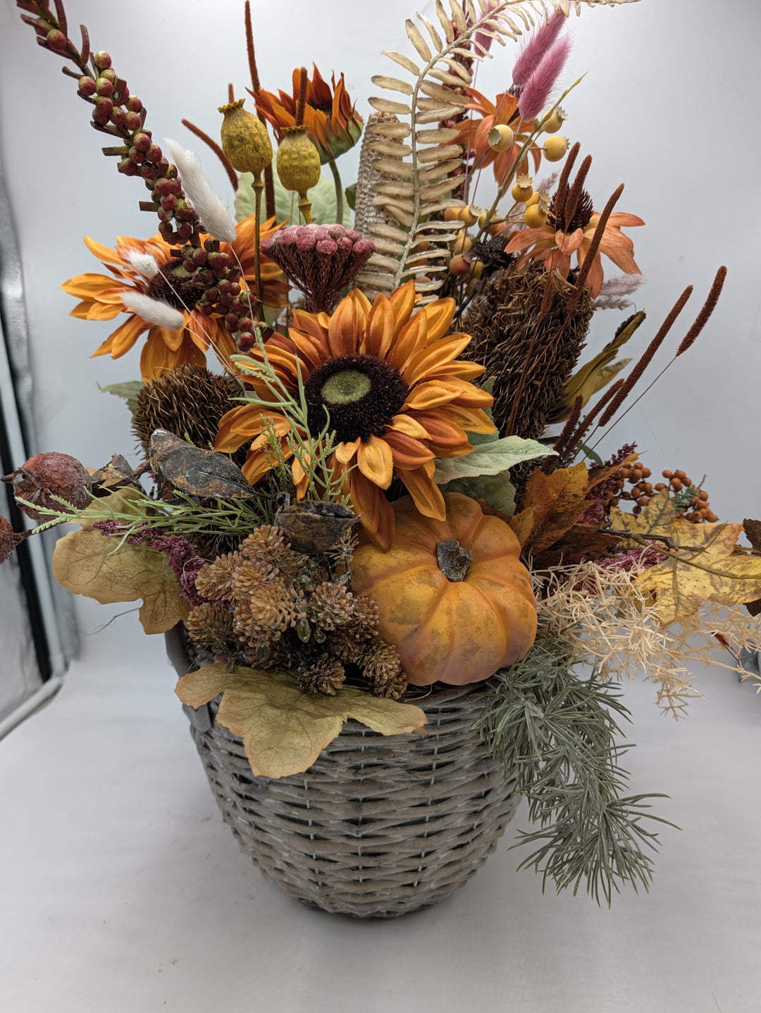 Hohes Herbstgesteck mit orangefarbenen Sonnenblumen, Kürbis, Zapfen und Gräsern in grauem Weidenkorb