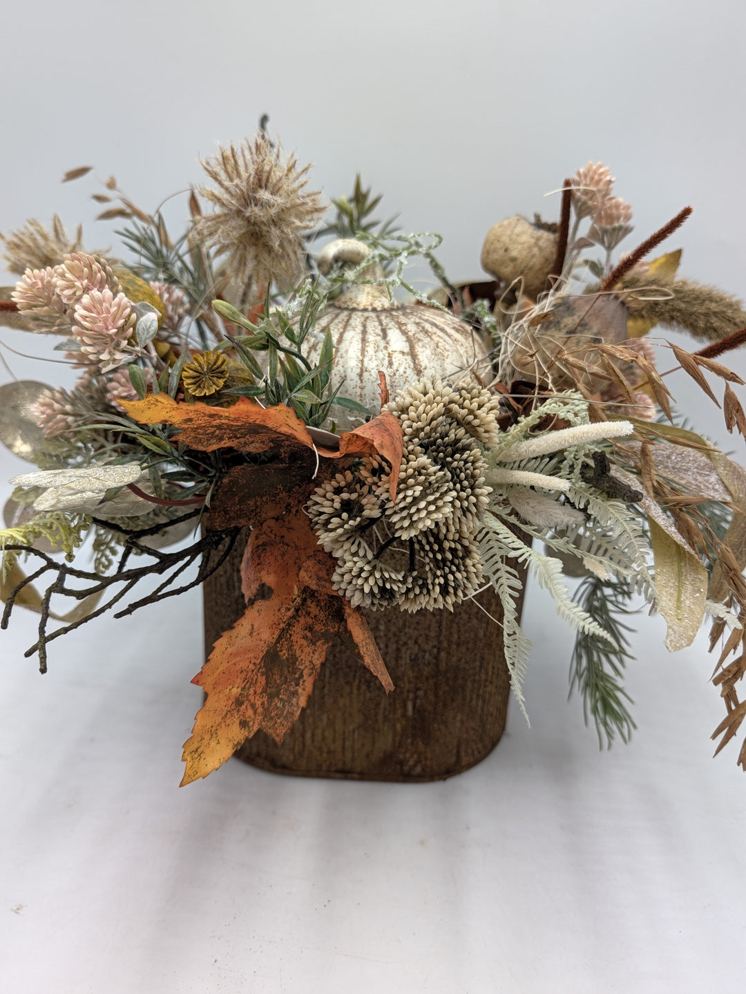 Herbstgesteck von vorn: cremefarbene Blütenbällchen, rosa Blüten, Farn und Gräser rund um einen Kürbis