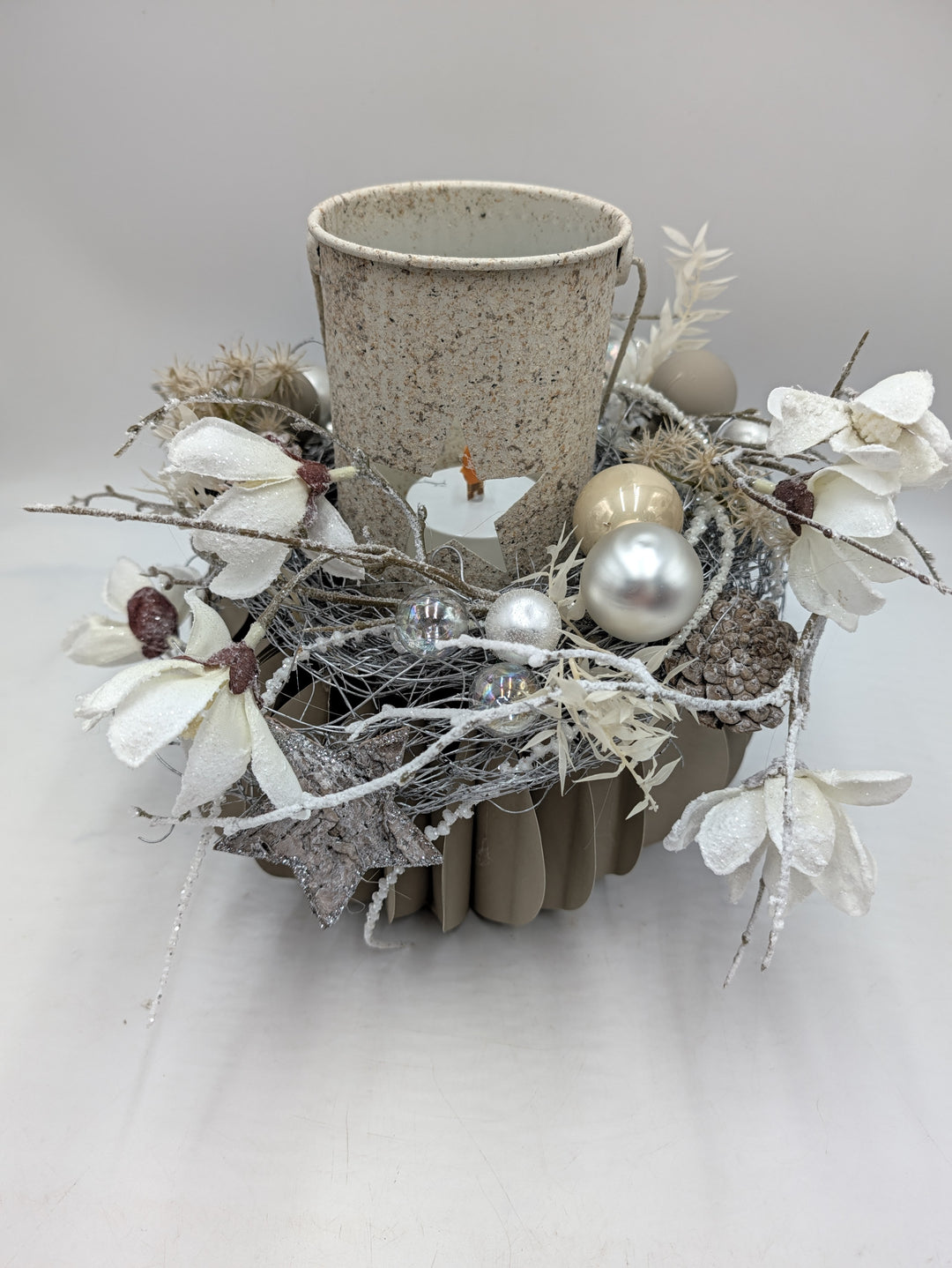 Wintergesteck von oben: Windlicht mit Kerze, beschneite Magnolienblüten, Zapfen und Kugeln in Silber und Creme