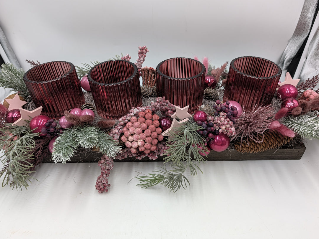 Adventsgesteck von vorne: rosa Beerenkugel, Holzsterne, Zapfen und dunkellila Trauben zwischen geriffelten Gläsern