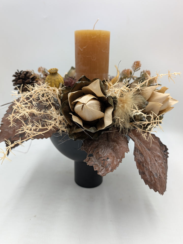 Herbstgesteck mit ockerfarbener Stumpenkerze, cremefarbener Blüte, Zapfen und braunen Blättern in schwarzer Schale