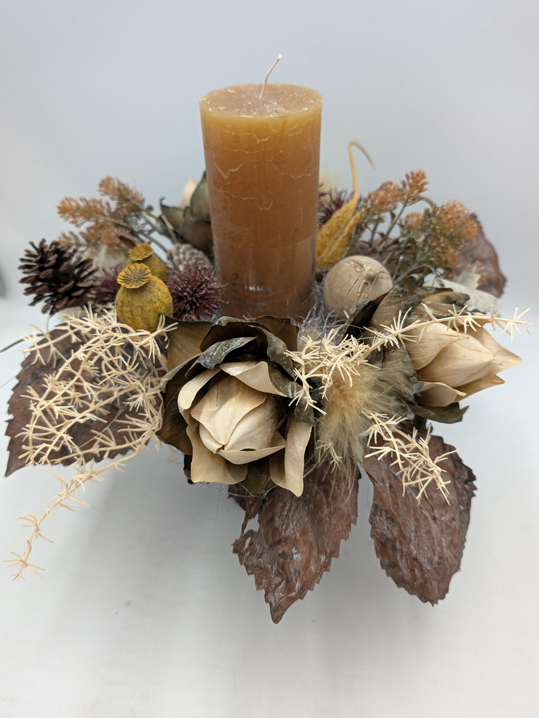 Herbstgesteck von oben: Kerze in Ocker, Mohnkapseln, Disteln, helle Gräser und breite braune Blätter
