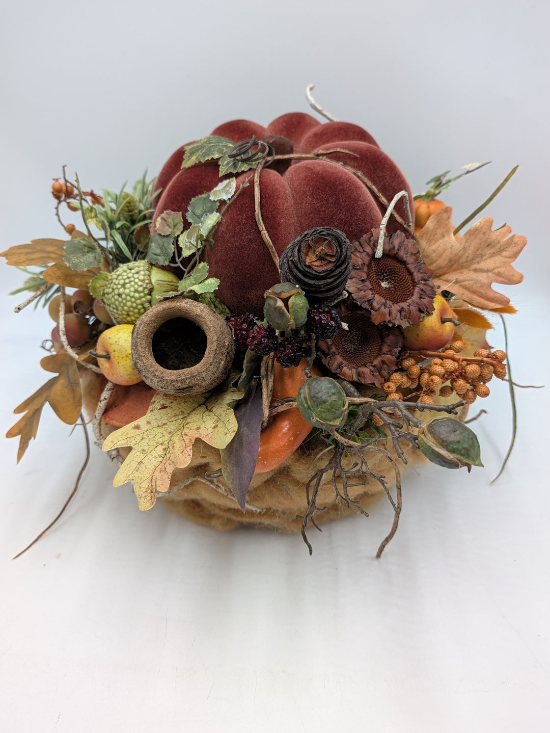 Herbstgesteck von vorn: weinroter Samtkürbis auf braunem Sockel, umgeben von Laub, Äpfeln, Beeren und Ranken