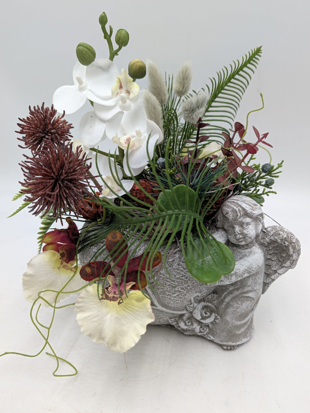 Graues Engel-Gefäß mit Orchideen, Gräsern, Kiefernzweigen und herabhängenden grünen Ranken, seitlich fotografiert