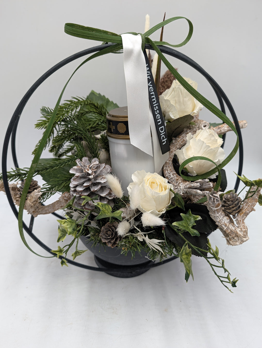 Nahsicht auf Grablicht mit goldenem Deckel, weiße Rosen, heller Zapfen, Tannenzweige und Satinband