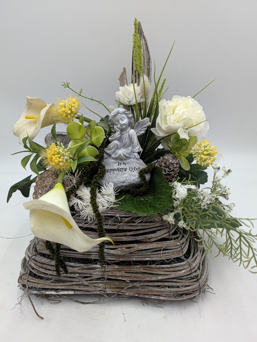 Trauergesteck mit Engelfigur, weißen Callas, Rose und gelben Blüten in eckigem Korb aus hellem Geflecht