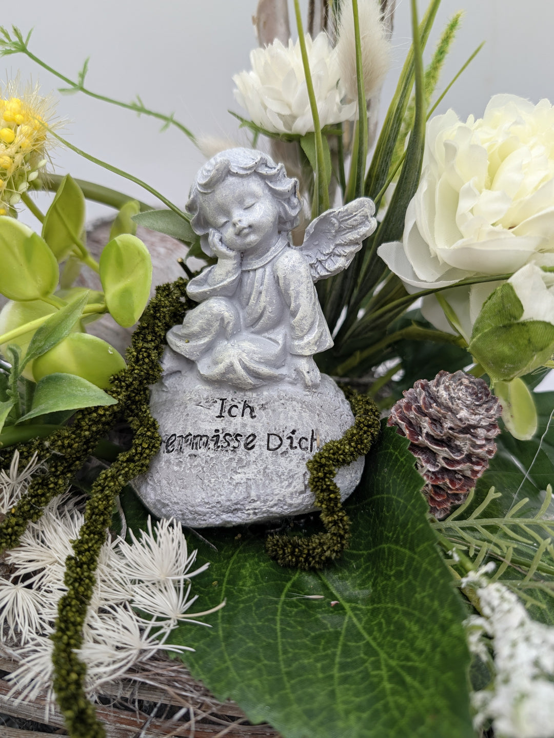 Nahaufnahme der silbergrauen Engelfigur auf einem Stein mit der Aufschrift Ich vermisse Dich, umgeben von Blüten und Moos