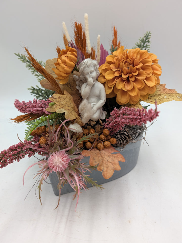 Trauergesteck mit weißer Engelsfigur, orangen Dahlien, rosa Heide und Eicheln in grauem eckigem Gefäß