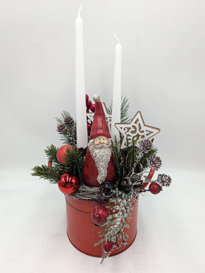 Weihnachtsgesteck im roten Metalltopf mit Weihnachtsmannfigur, zwei weißen Stabkerzen, Tannenzweigen und Holzstern