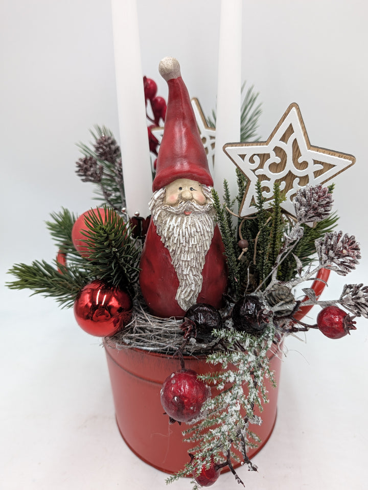 Nahaufnahme des Gestecks: Weihnachtsmann mit weißem Bart, rote Kugeln, Hagebutten, beschneite Zapfen und Holzstern