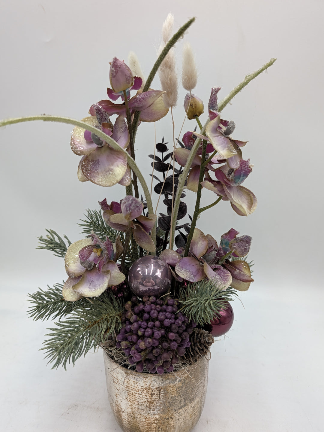 Gesteck von vorn: Orchideenblüten, violette Beerendolde, flieder Kugeln und silbrige Tannenzweige im Goldtopf
