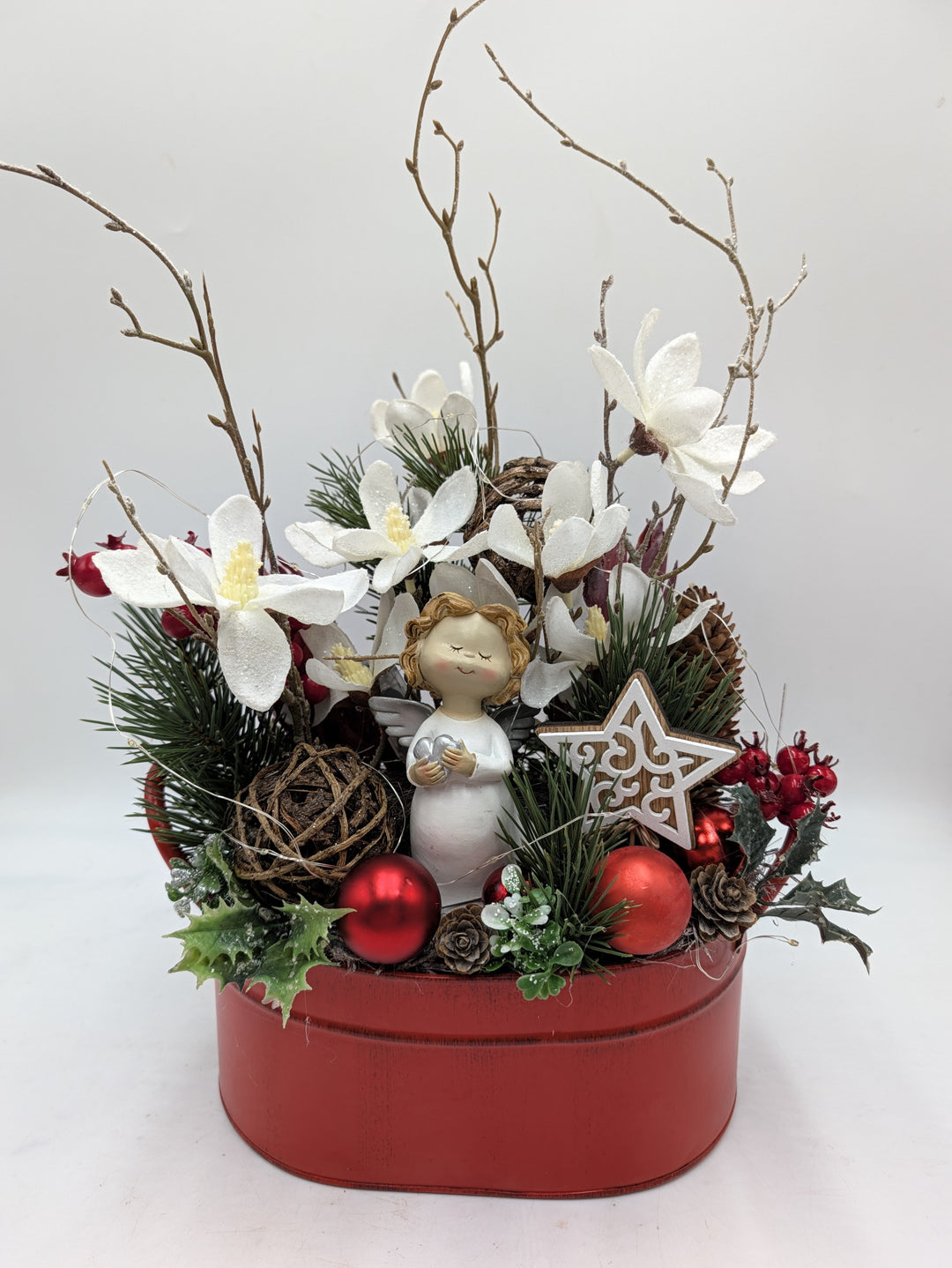 Weihnachtsgesteck mit Engelfigur, weißen Magnolien, roten Kugeln und Zweigen in roter Metallschale